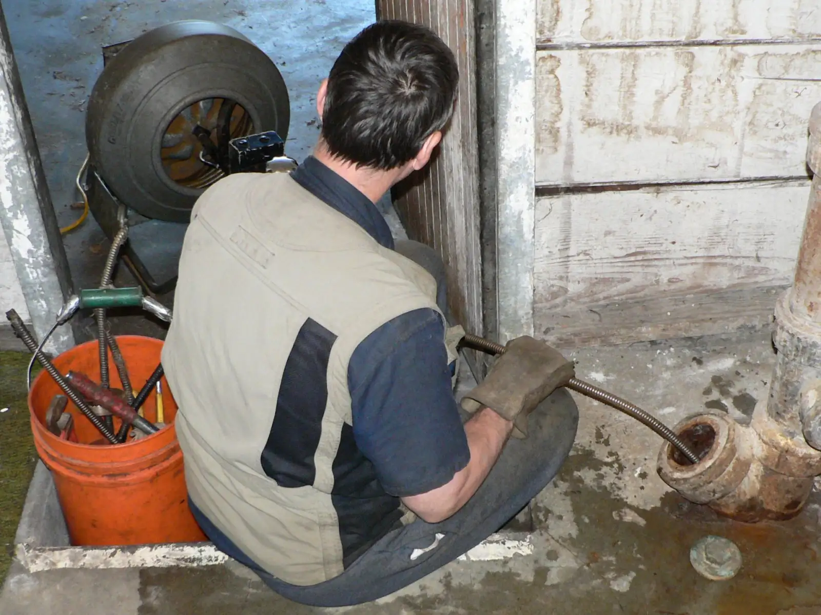 Homme vu de dos, assis au sol dans un sous-sol, guidant d'une main gantée le câble d'un furet motorisé à tambour dans l'ouverture d'une canalisation en fonte