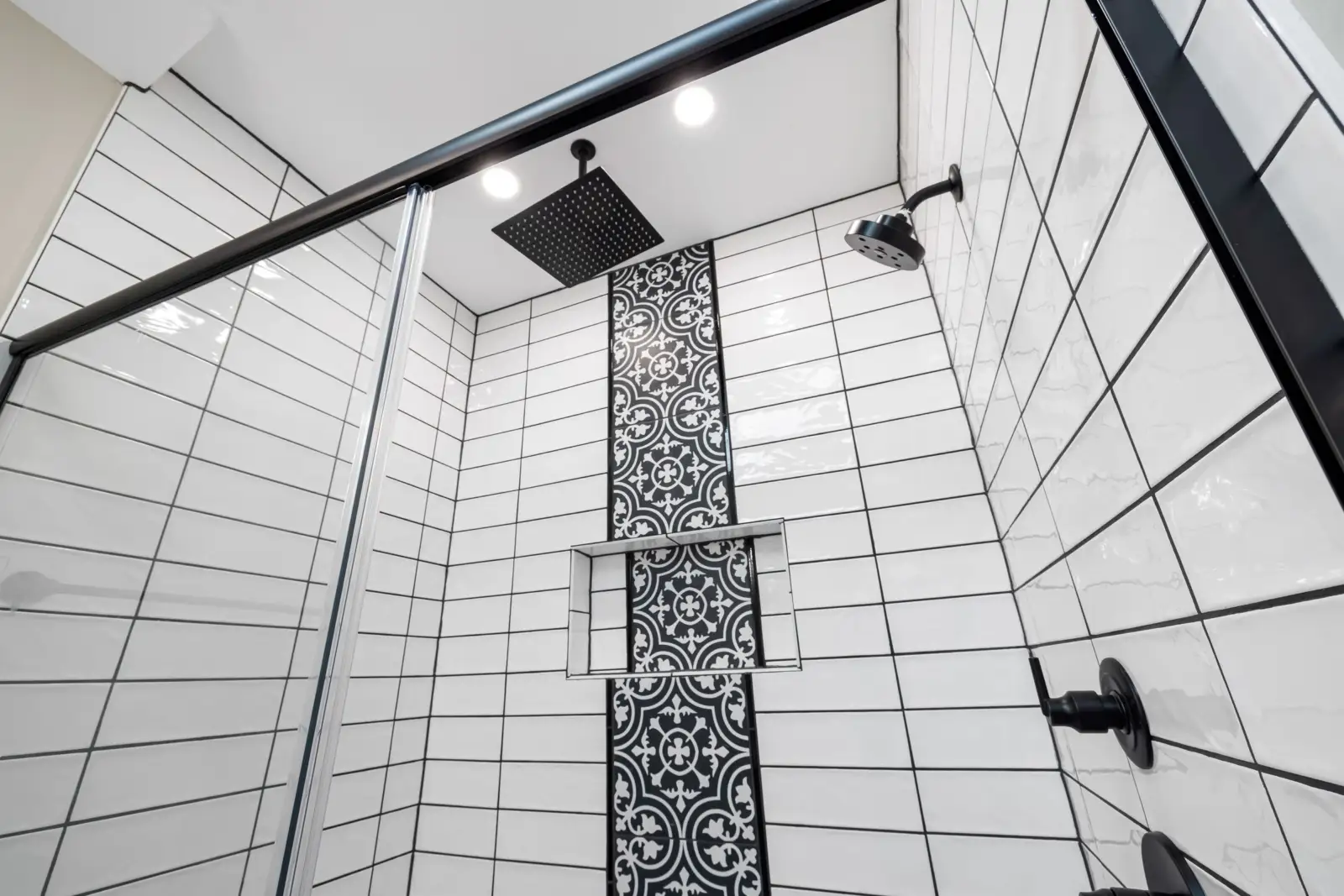 Cabine de douche carrelée de blanc avec un ciel de pluie carré noir au plafond et un pommeau orientable fixé au mur