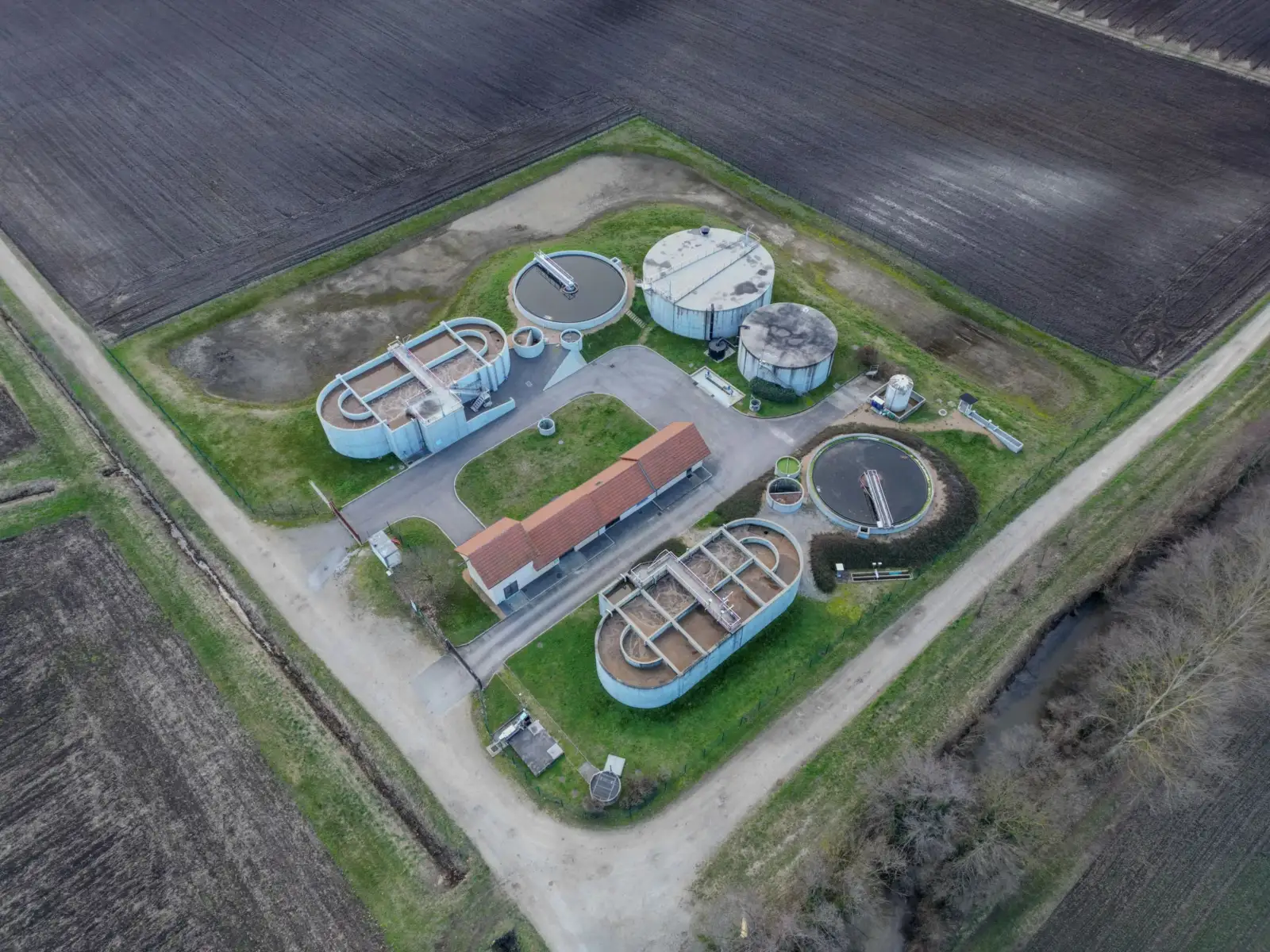 Vue aérienne d'une petite station d'épuration entourée de champs labourés, avec ses bassins circulaires et un bâtiment à toit de tuiles