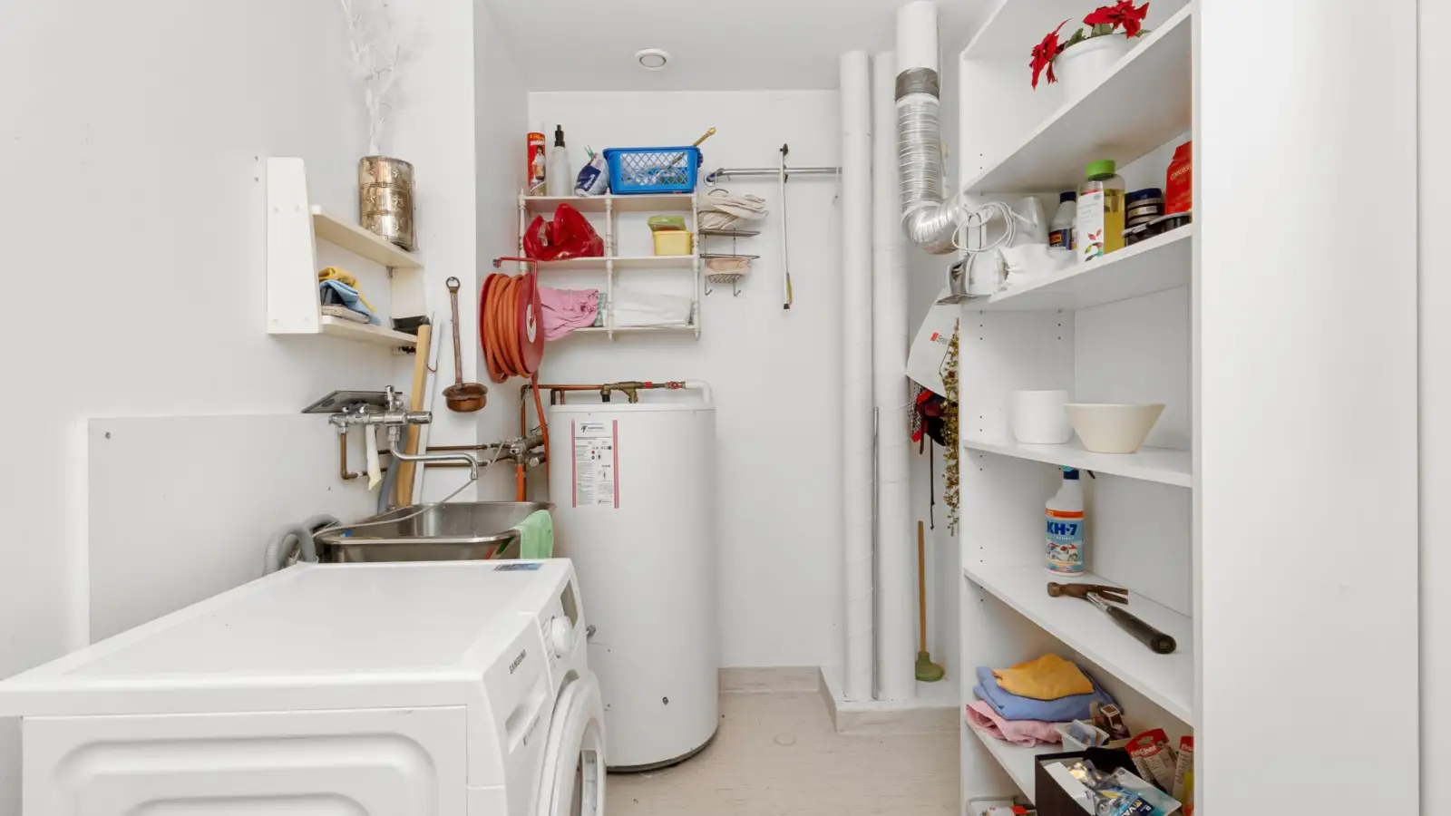 Chauffe-eau électrique vertical posé au sol dans une buanderie, entre un évier, des étagères et un lave-linge