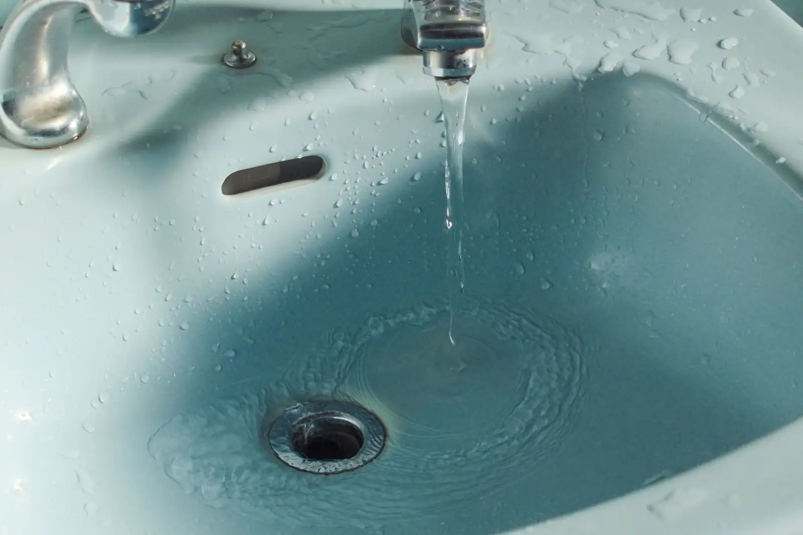 Filet d'eau qui coule d'un robinet chromé dans un lavabo bleu clair et tourbillonne vers la bonde