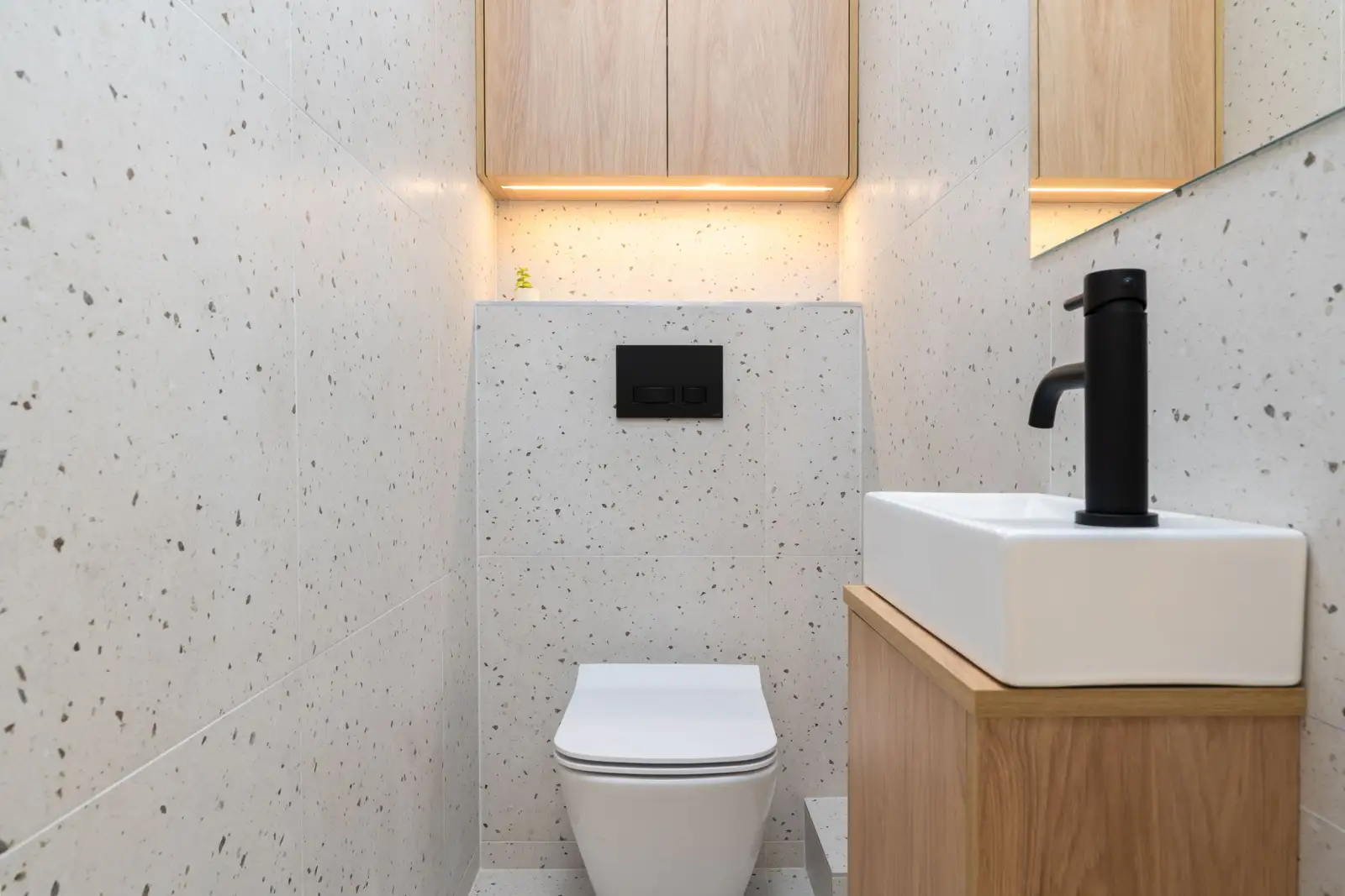 Toilettes étroites avec WC suspendu blanc, coffrage carrelé à mi-hauteur, plaque de commande noire et petit lave-mains sur meuble en bois