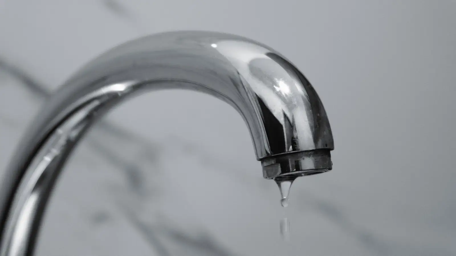 Bec de robinet chromé d'où tombe une goutte d'eau, devant un mur en marbre gris