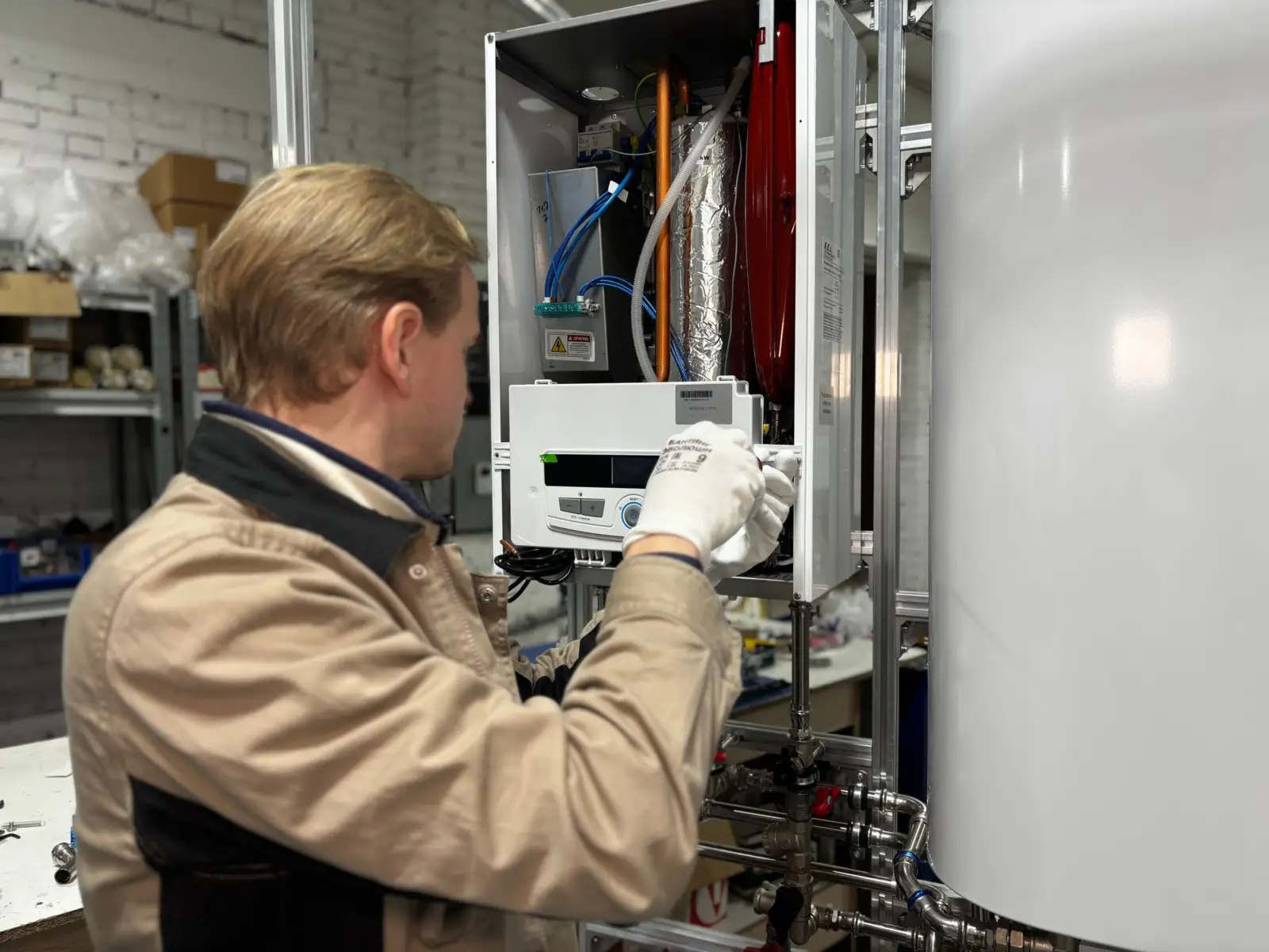 Technicien de dos réglant une chaudière murale capot ouvert, à côté d'un grand ballon de stockage blanc, dans un atelier