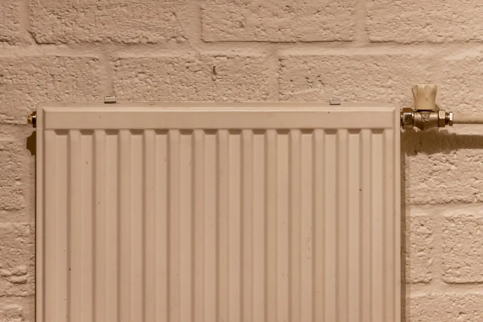 Radiateur en acier blanc fixé sur un mur de briques peint, avec un robinet manuel à droite et un bouchon de purge en haut à gauche