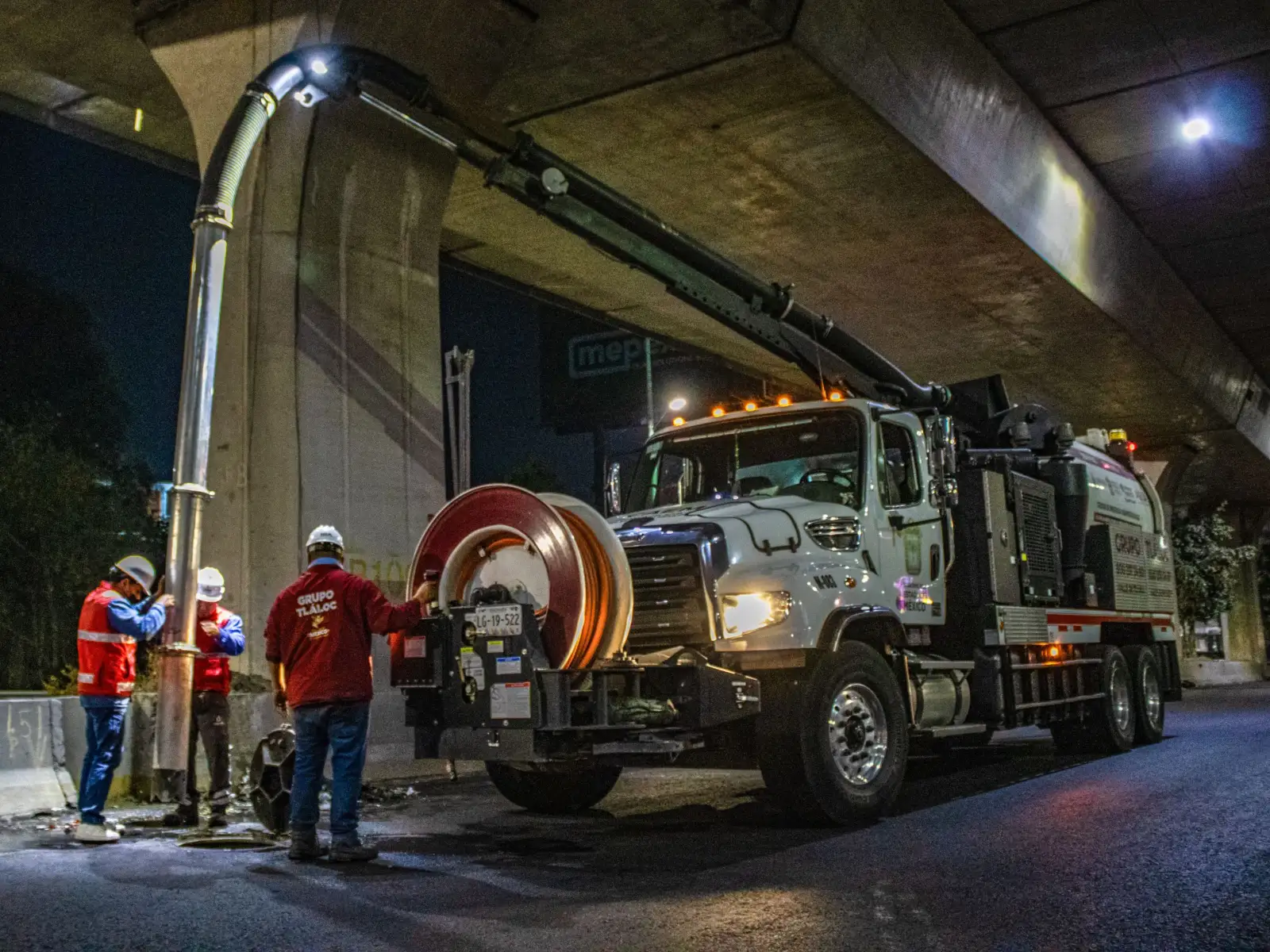 Camion aspirateur hydrocureur en intervention de nuit sous un pont routier, flexible d'aspiration déployé et agents en gilet