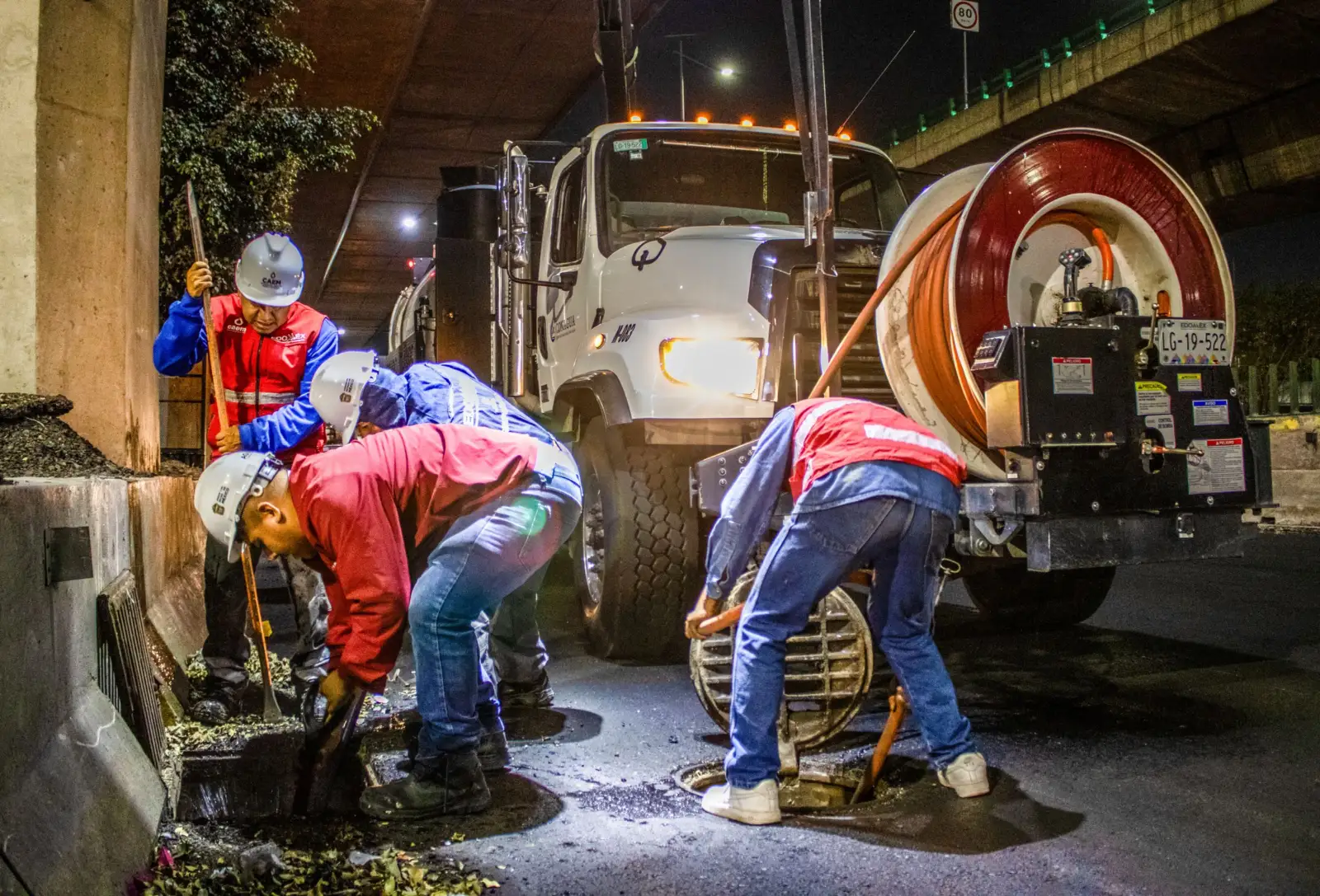 Camion hydrocureur de nuit avec son grand enrouleur de flexible, et des agents casqués penchés sur un regard de voirie ouvert