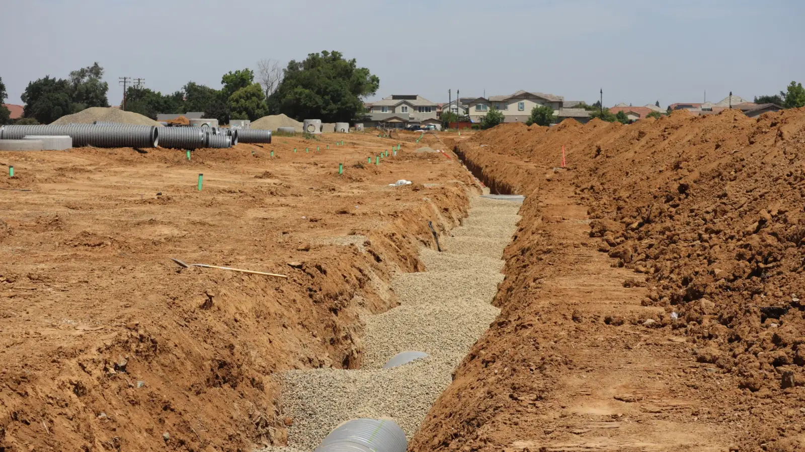 Tranchée ouverte sur un chantier de lotissement, fond garni de gravier avec un tuyau posé au fond, tubes annelés stockés à l'arrière-plan