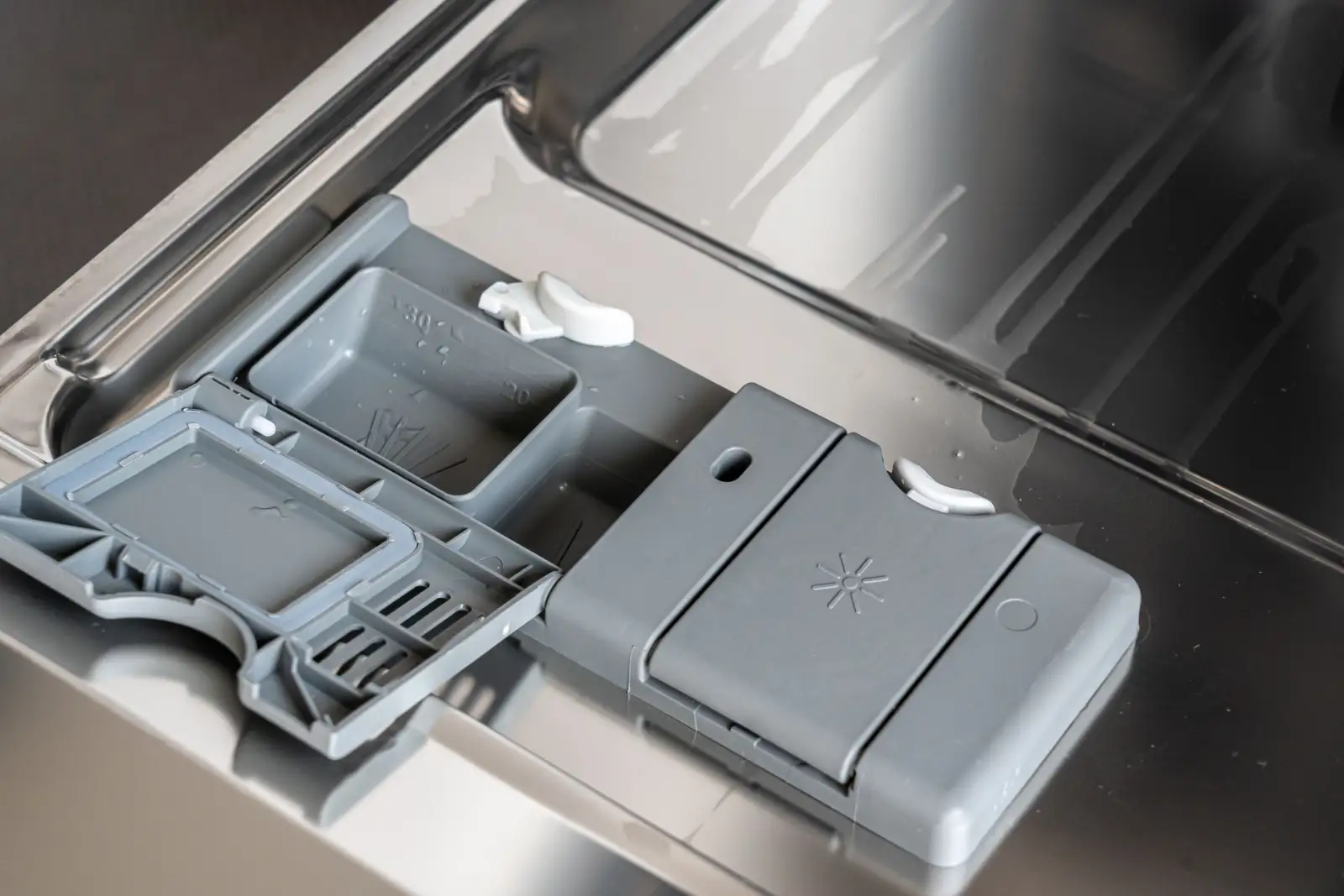 Bac à produit de lavage gris ouvert sur la contre-porte en inox d'un lave-vaisselle, avec son couvercle rabattu et le réservoir de liquide de rinçage