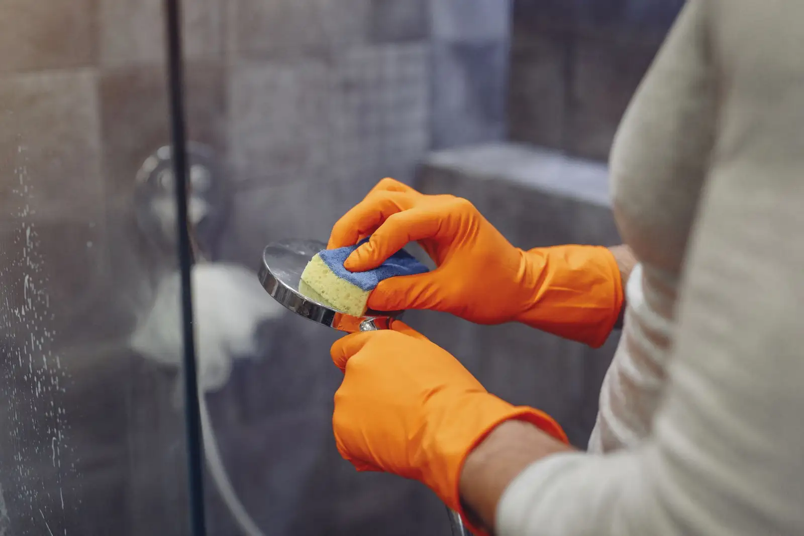 Mains gantées d'orange nettoyant un pommeau de douche avec une éponge