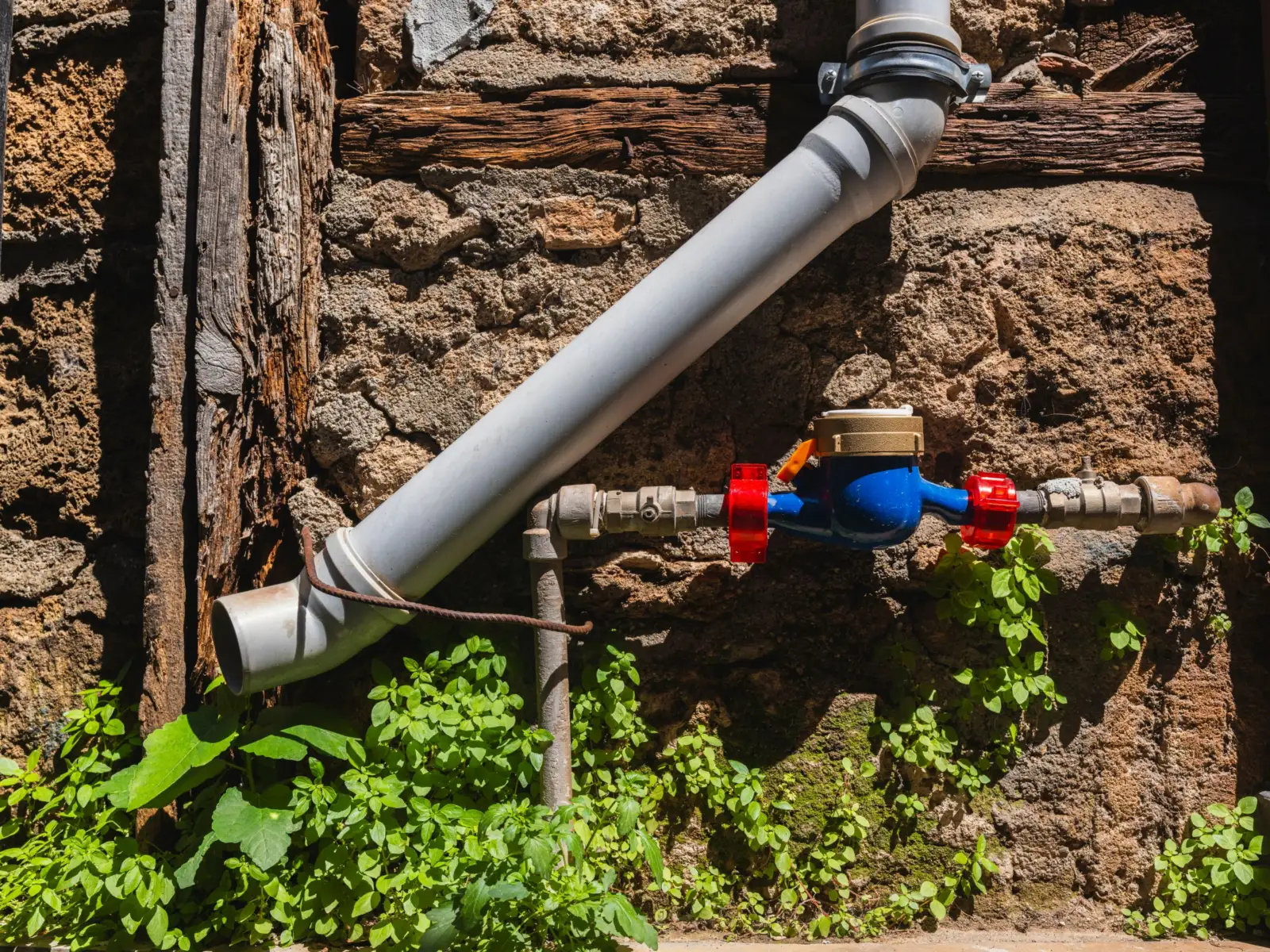 Compteur d'eau à corps bleu et raccords rouges monté sur un tuyau le long d'un mur de pierre, à côté d'un tuyau d'évacuation gris