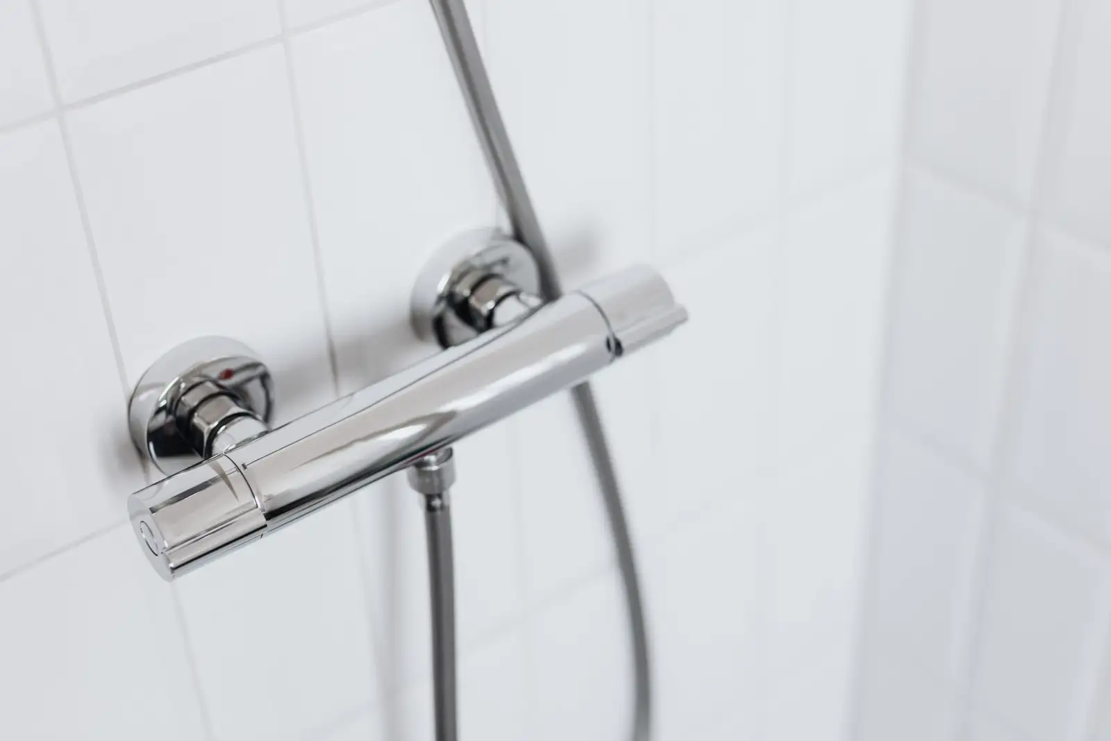 Mitigeur thermostatique chromé fixé sur deux raccords muraux, surmonté du tube d'une colonne de douche et d'un flexible, sur carrelage blanc