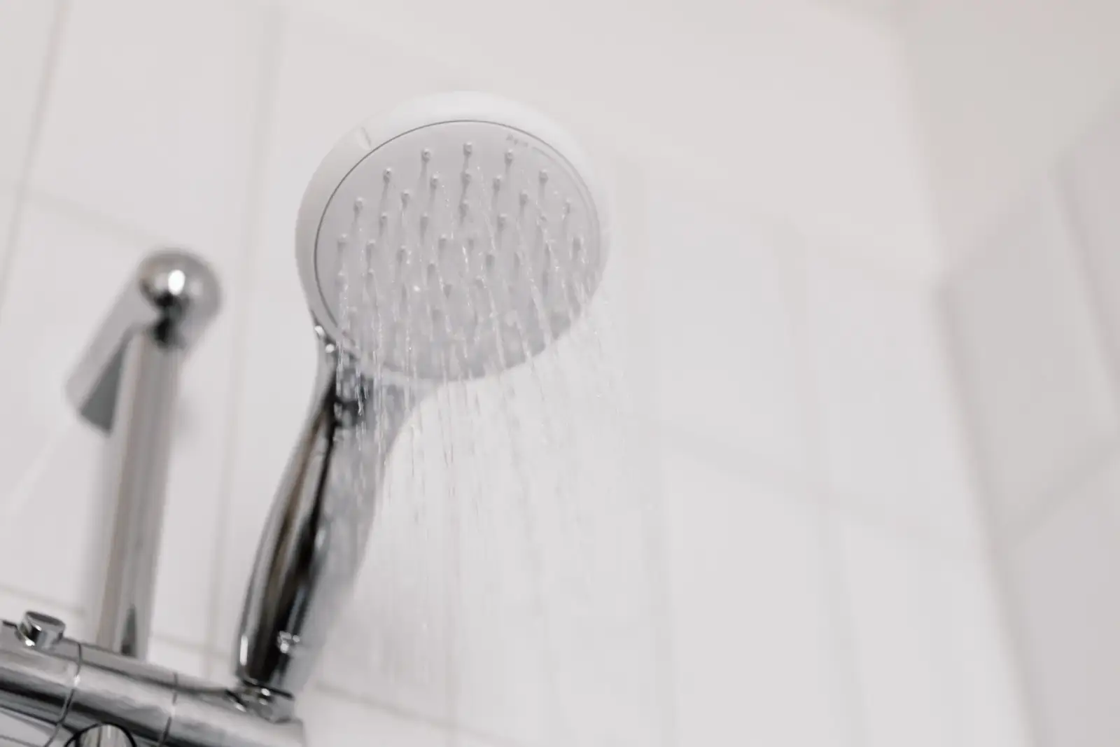 Douchette à tête blanche et manche chromé posée sur sa barre, l'eau s'écoulant en fins filets devant un carrelage blanc