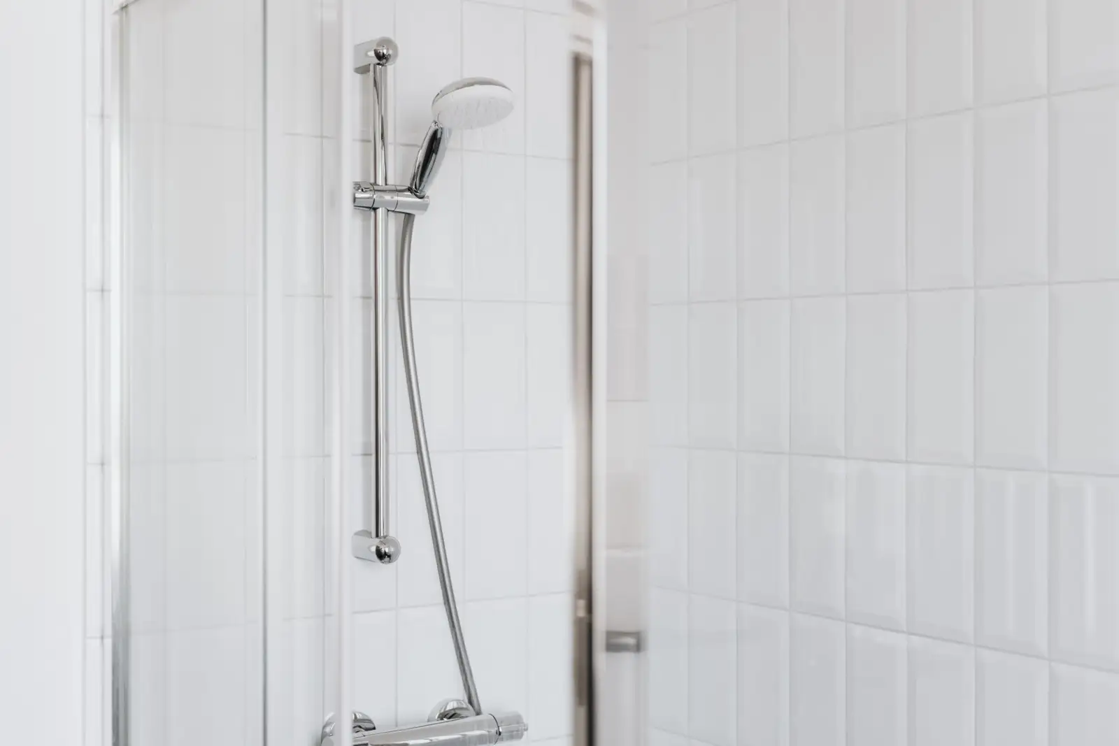 Barre de douche chromée avec douchette blanche posée sur son support et flexible descendant vers le mitigeur, devant un carrelage blanc