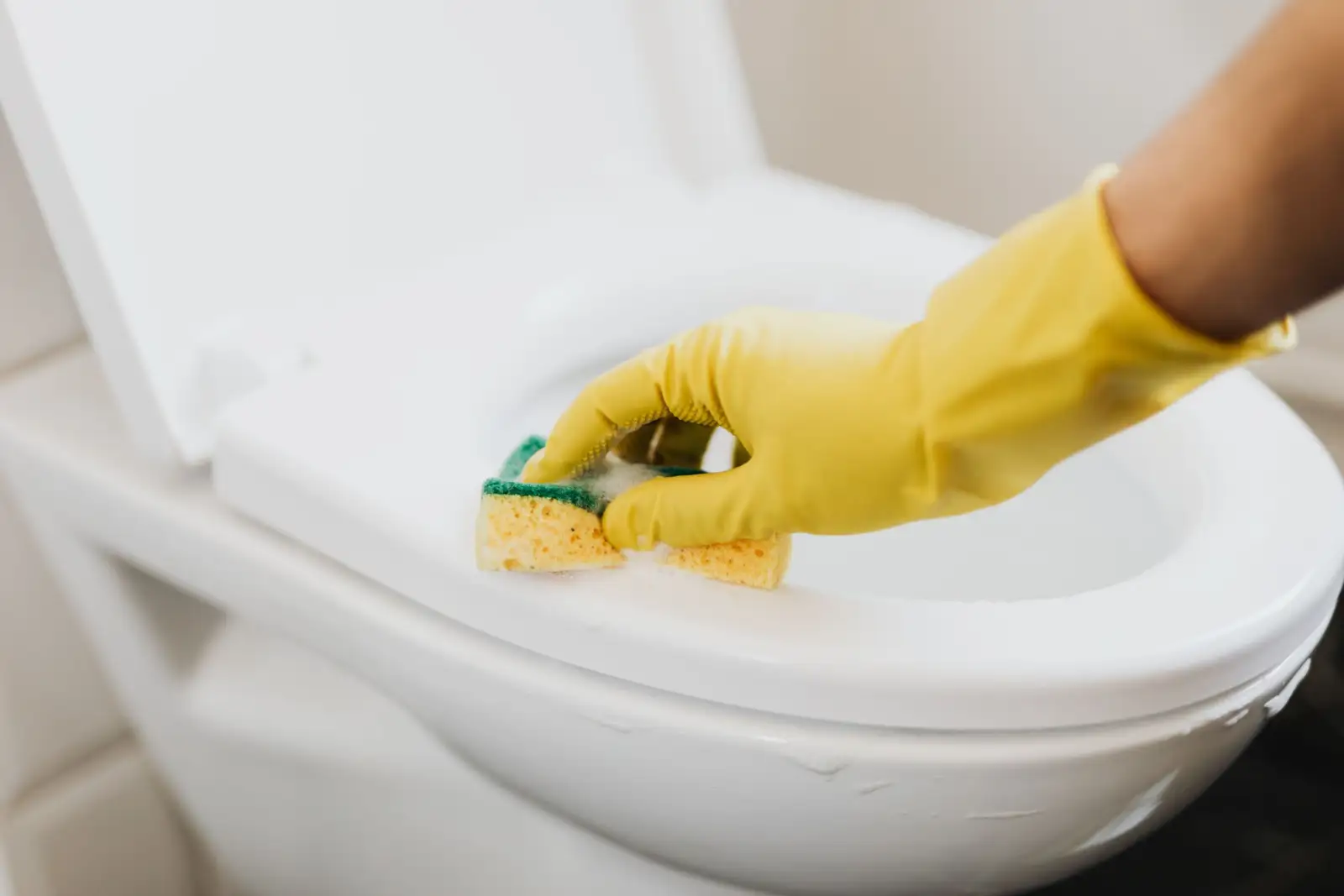 Main gantée de jaune qui nettoie à l'éponge le rebord et l'abattant d'une cuvette de WC blanche