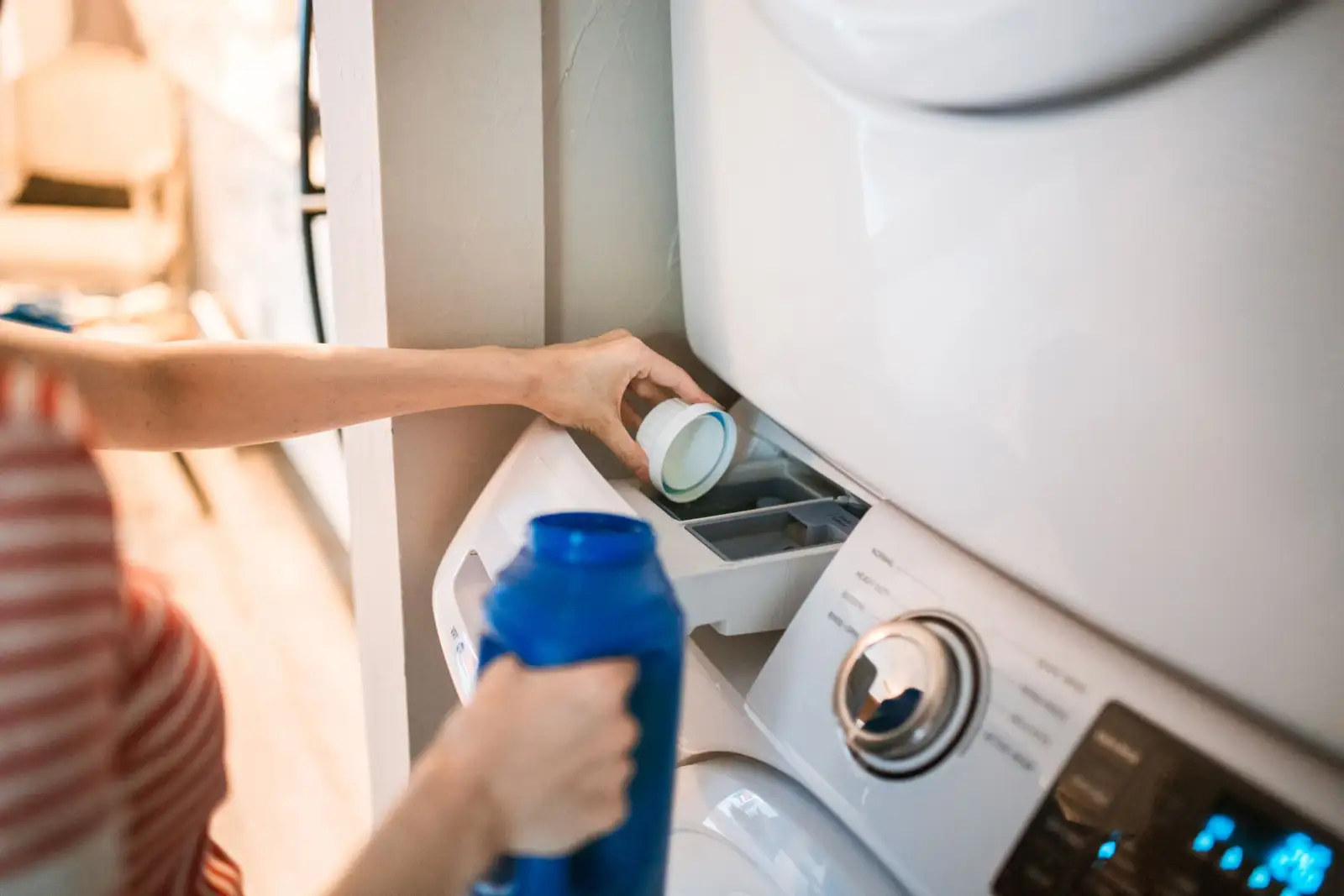 Main qui vide un bouchon doseur de lessive dans le bac à produits d'un lave-linge, un flacon bleu dans l'autre main