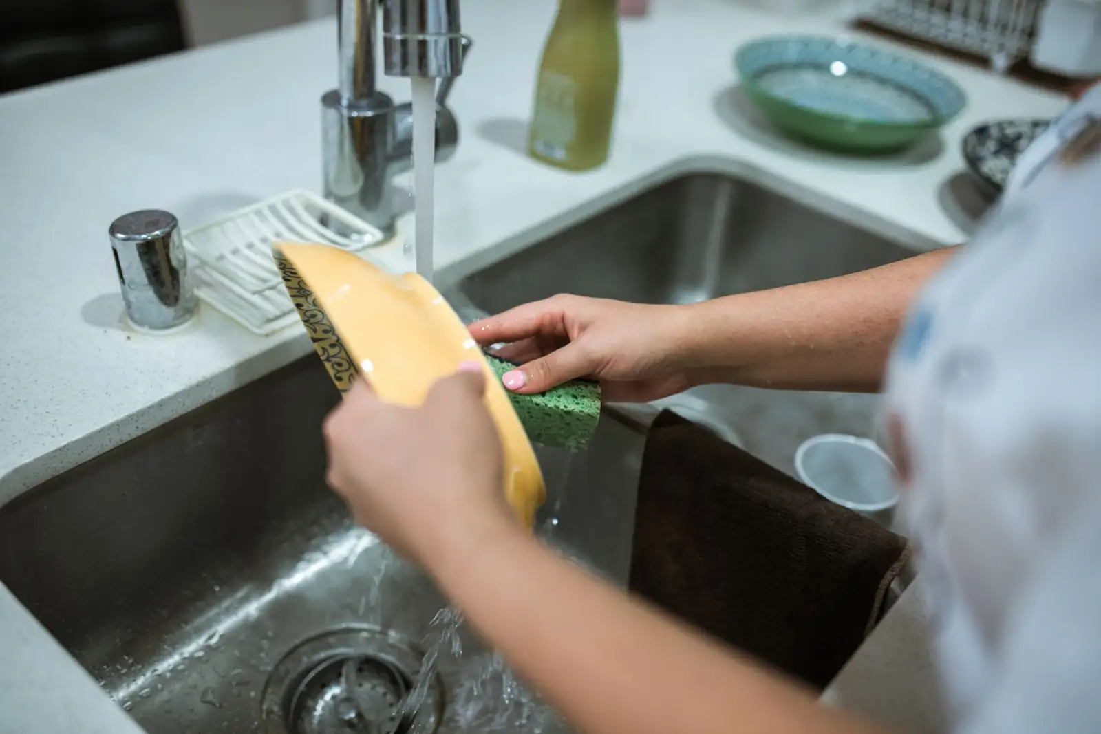 Mains lavant une assiette sous le robinet d'un évier de cuisine en inox, l'eau coulant vers la bonde