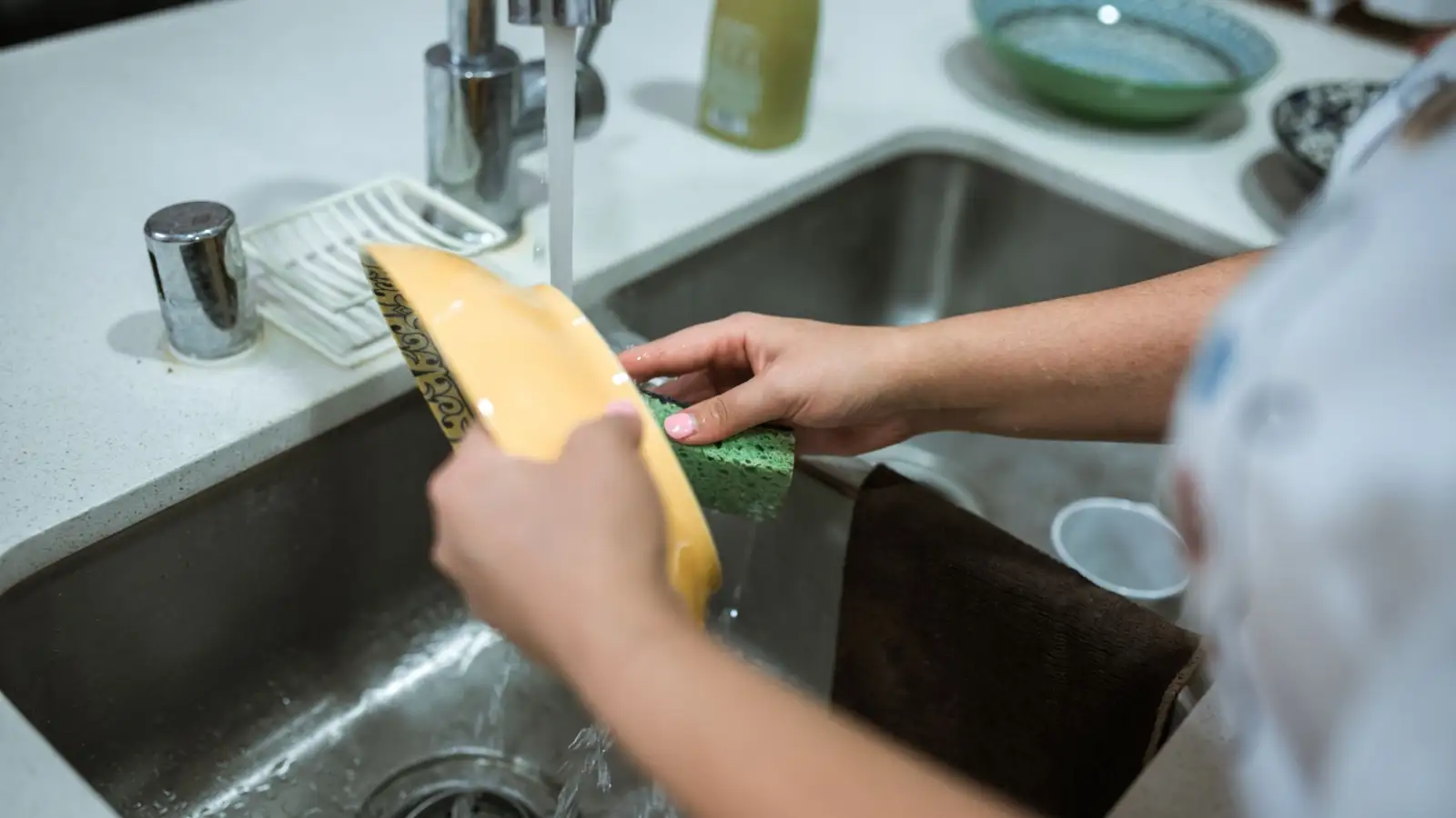 Mains lavant une assiette sous le robinet d'un évier de cuisine en inox, l'eau coulant vers la bonde