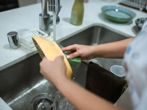 Mains lavant une assiette sous le robinet d'un évier de cuisine en inox, l'eau coulant vers la bonde