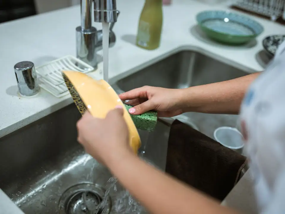 Mains lavant une assiette sous le robinet d'un évier de cuisine en inox, l'eau coulant vers la bonde