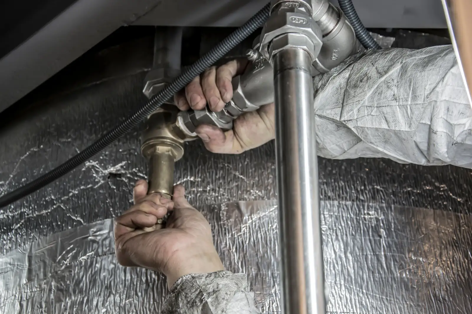 Deux mains serrant un raccord en laiton sur une canalisation métallique, devant un isolant aluminium