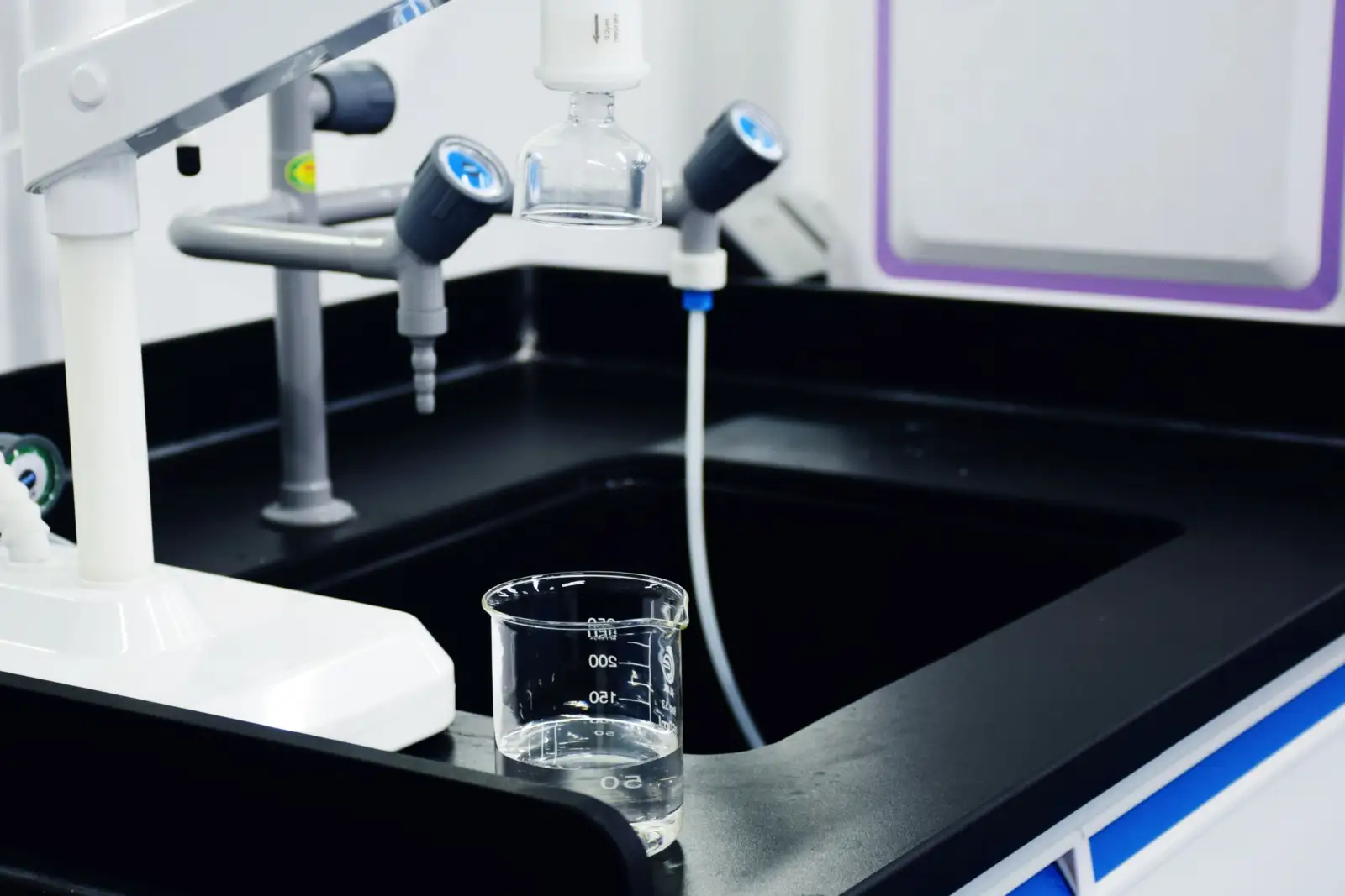 Bécher gradué en verre rempli d'eau posé au bord d'un évier de paillasse de laboratoire, sous des robinets de laboratoire
