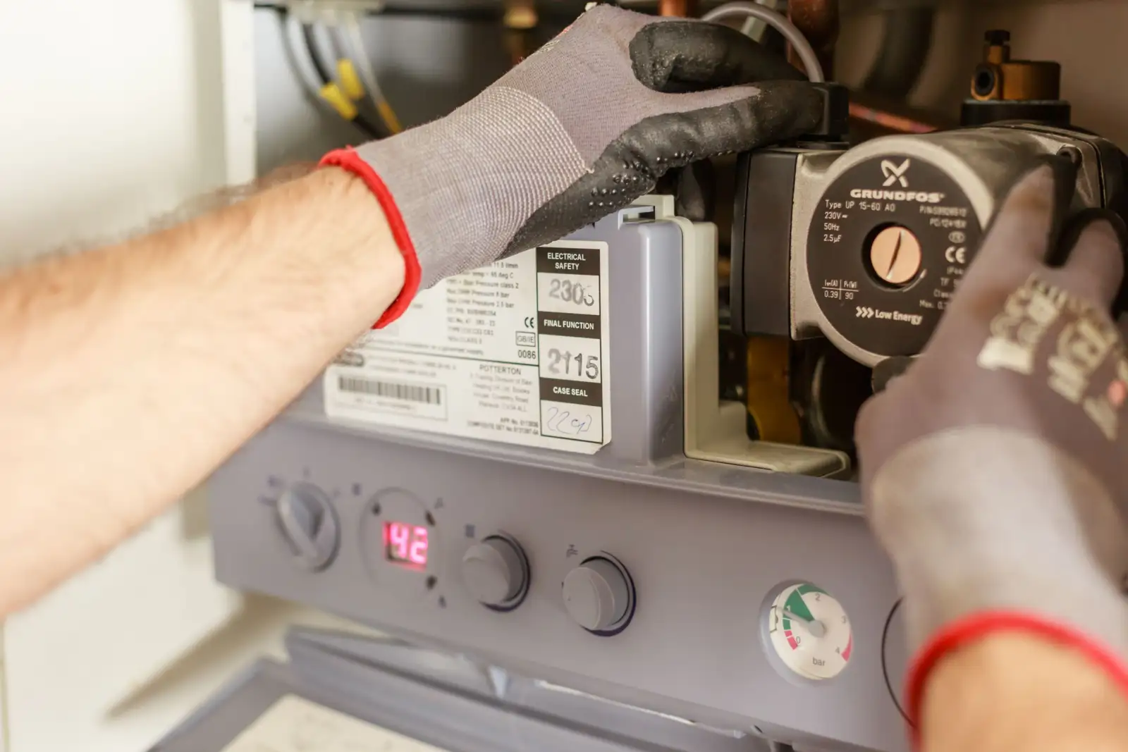 Mains gantées d'un technicien sur une chaudière murale ouverte, avec le circulateur, le panneau de commande et le manomètre visibles